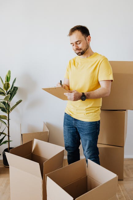 A man wearing a yellow T-shirt and blue jeans stands indoors, holding a clipboard and pen, inspecting or planning the packing process during a home relocation. Surrounding him are open cardboard boxes