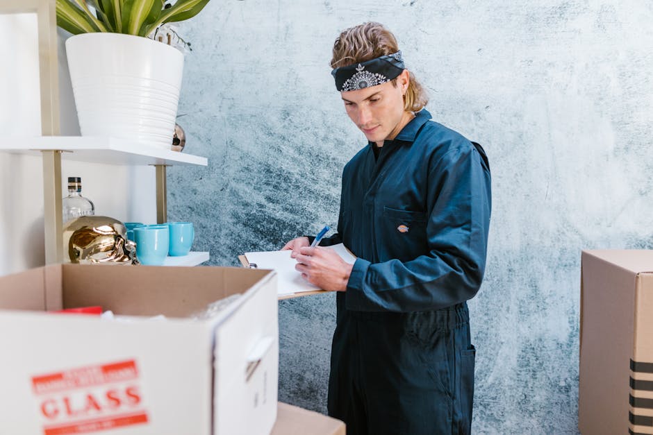 A man wearing a yellow T-shirt and blue jeans stands indoors, holding a clipboard and pen, inspecting or planning the packing process during a home relocation. Surrounding him are open cardboard boxes
