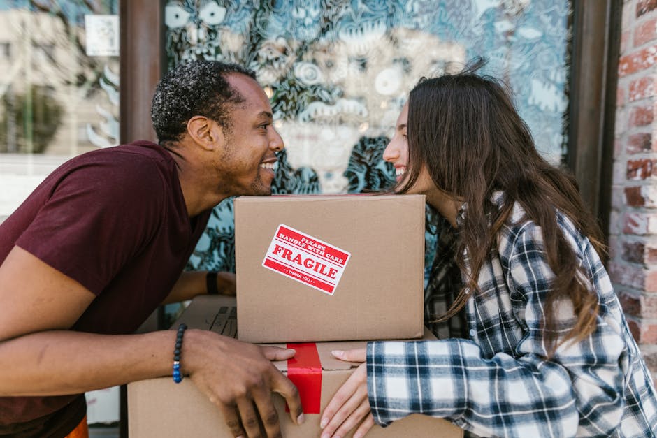 A man and woman, both smiling, face each other closely while holding and embracing a cardboard box with red and white 'Fragile' handling labels during a house move. The man, with dark curly hair and w