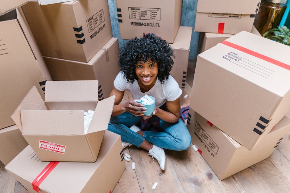 A man and woman, both smiling, face each other closely while holding and embracing a cardboard box with red and white 'Fragile' handling labels during a house move. The man, with dark curly hair and w