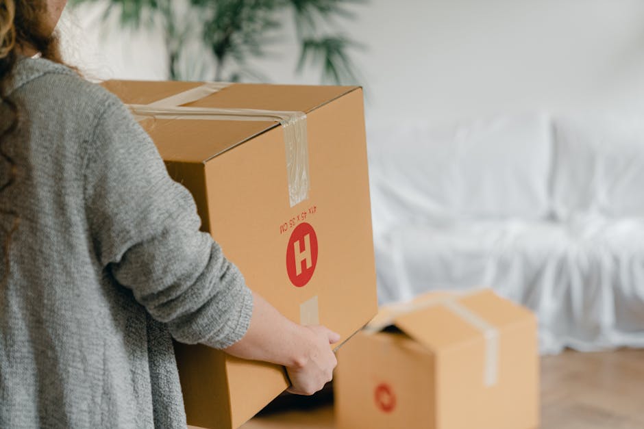 A person wearing a grey knitted sweater is holding a medium-sized cardboard box sealed with clear packing tape, with a red logo and printed text, preparing to load it into a moving van. Several other cardboard boxes of various sizes, also sealed with tape, are positioned nearby on the floor inside a room featuring light wooden flooring and a white sofa covered with a slipcover in the background. The scene depicts the process of packing and home relocation, with boxes ready for furniture transport and moving logistics, aligning with professional removals services offered by Man With a Van Hillingdon. The lighting is natural, casting soft shadows, and the environment suggests an indoor setting being prepared for a house move or packing and moving operation.