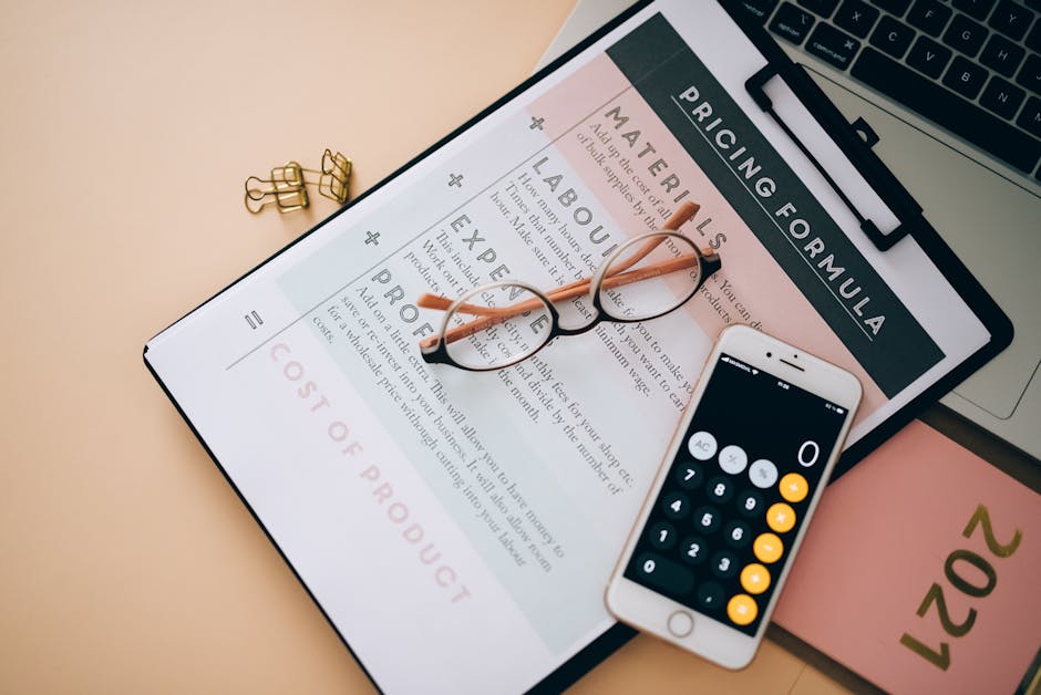A close-up view of a paper document titled 'Pricing Formula' displaying sections for materials, labour, expense, and profit along with a large red price figure of £350 at the bottom, resting on a light-colored surface. On top of the document, there are a pair of black-rimmed glasses with orange temples, a white smartphone with a calculator app open displaying the number 0, a few small gold binder clips, and part of a silver laptop with a black keyboard visible in the background. The setting appears to be an indoor workspace related to house removals, packing, or moving logistics, with the materials and devices suggesting planning or cost estimation activities often involved in home relocation services provided by Man With a Van Hillingdon.