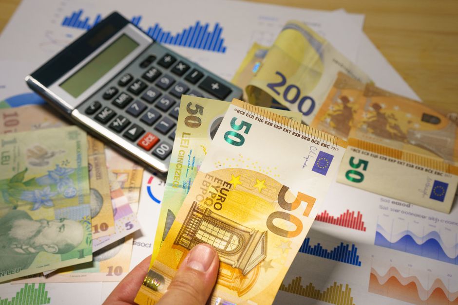 A close-up view of various euro banknotes, including €50, €20, and €10 denominations, being held by a person over a desk with financial documents, charts, and a calculator. The banknotes are spread out, with some partially overlapping each other, and are positioned near a small, black calculator that is turned on. In the background, there are printed sheets displaying bar graphs and line charts in red, blue, green, and orange, suggesting financial analysis related to home removals or moving costs. The scene reflects the financial considerations involved in house removals, such as cost estimation and budgeting, aligning with the services offered by Man With a Van Hillingdon for home relocation and furniture transport, and supporting the topic of how removals pricing is calculated.