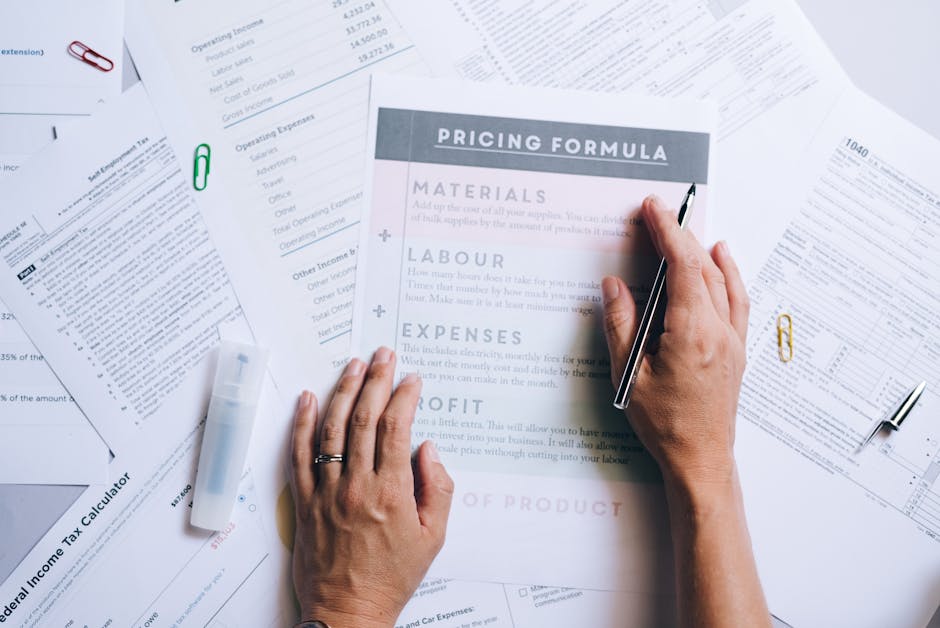A close-up view of a paper document titled 'Pricing Formula' displaying sections for materials, labour, expense, and profit along with a large red price figure of £350 at the bottom, resting on a light-colored surface. On top of the document, there are a pair of black-rimmed glasses with orange temples, a white smartphone with a calculator app open displaying the number 0, a few small gold binder clips, and part of a silver laptop with a black keyboard visible in the background. The setting appears to be an indoor workspace related to house removals, packing, or moving logistics, with the materials and devices suggesting planning or cost estimation activities often involved in home relocation services provided by Man With a Van Hillingdon.