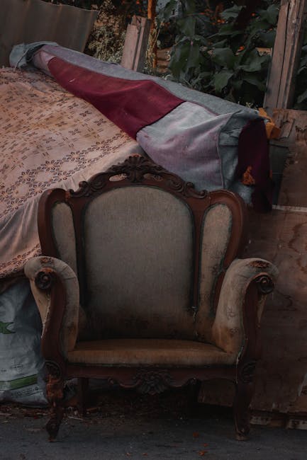 A vintage armchair with a wooden frame and upholstered fabric seat and backrest, positioned indoors on a wooden floor. Behind the chair, a rolled-up carpet with patterned fabric and red, grey, and beige sections is partially draped over a surface, possibly a table or another piece of furniture. To the left, a plastic bag is visible on the floor, and in the background, there are plants with large green leaves and a wooden structure, suggesting the setting may be a garage or storage area. The lighting appears natural, indicating daylight, and the environment looks organized for a home relocation or furniture moving process, with items in temporary storage before being transported by Man With a Van Hillingdon for their removals service.