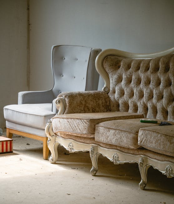 Inside a room with plain, light-colored walls and a concrete or unfinished floor, two vintage armchairs are positioned near a wall. The first armchair on the left is upholstered in light fabric with button-tufted detailing on the backrest and wooden legs, while the second armchair on the right is an ornate Victorian-style piece with a carved wooden frame and plush, velvet-like fabric in a beige tone, featuring tufted upholstery on the backrest and arms. A small dark object, possibly a remote or a tool, rests on the seat of the second chair. The setting appears to be part of a home undergoing furniture removal or relocation, supported by [COMPANY_NAME], which specialises in removals and home relocation services. This scene depicts a typical stage in furniture transport, with no other objects such as boxes or packaging materials visible, indicating that the focus is on the furniture ready for moving.