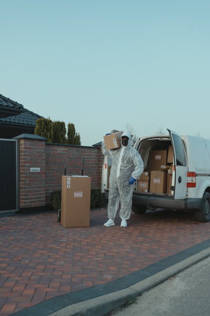 A person dressed in a full white protective suit, including gloves and a hood, is standing outdoors on a paved driveway next to an open white van used for house removals. The individual is holding a cardboard box, ready to load or unload it from the vehicle. Several other cardboard boxes, some sealed with packing tape, are placed on the ground nearby. The van's interior is visible, filled with more packed boxes of various sizes, indicating an ongoing house relocation process. The van is parked adjacent to a brick wall with a black gate, and in the background, there are trees and a clear blue sky, suggesting daytime. The scene is part of professional packing and furniture transport activities by Man With a Van Hillingdon, demonstrating typical steps involved in home relocation and moving services.