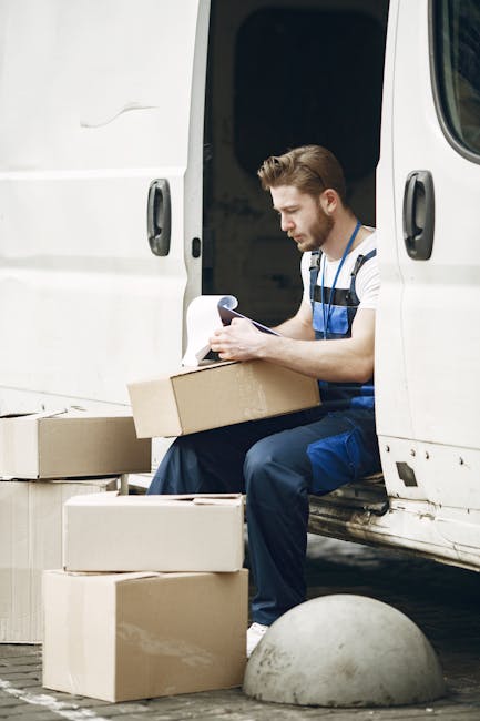 A young man with a beard and short brown hair, wearing a striped t-shirt and blue work overalls, is seated at the rear of a white van during a home relocation. He is holding a clipboard and appears to be reviewing documents as part of the packing and moving process. In front of him on the ground are several cardboard boxes of various sizes, some open and some closed, likely filled with household items prepared for transport. The van’s sliding door is open, revealing a dark interior. The scene is set outdoors, possibly on a driveway or pavement, with a concrete bollard visible nearby, indicating a residential or commercial property environment. The lighting suggests natural daylight, and the overall scene depicts a professional furniture transport and packing process, consistent with [COMPANY_NAME]’s house removals services in Hillingdon, focusing on efficient loading, documentation, and organized packing for a comprehensive home relocation.