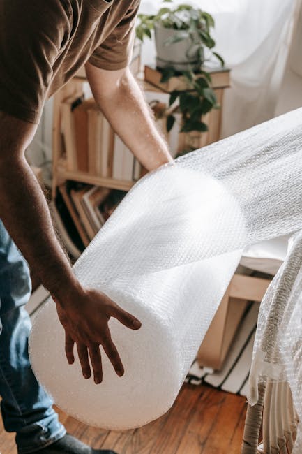 A person wearing a brown shirt is carefully unwrapping a large piece of white bubble wrap from a household item, likely during packing for a home relocation. The bubble wrap is partially unrolled and held with both hands, revealing the textured plastic surface designed for protecting furniture or appliances. The setting appears to be inside a well-lit room with wooden flooring, and visible background elements include a wooden bookshelf filled with books, a potted plant on a small stand, and a sheer white curtain allowing natural light to fill the space. This scene illustrates the careful packing process involved in home removals and furniture transport, highlighting the importance of secure wrapping during household moves coordinated by a professional service like Man With a Van Hillingdon, specializing in house removals and logistical moving solutions.
