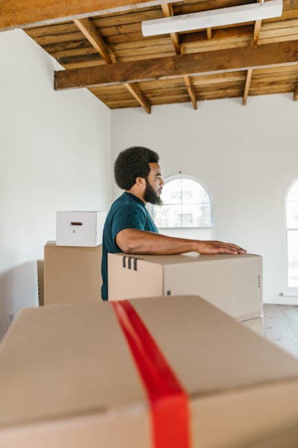 A man with a dark skin tone and curly hair, dressed in a dark green shirt, is inside a spacious room with a high, wooden-beamed ceiling and white-painted walls. He is standing behind a stack of packing boxes, holding onto one of the large cardboard boxes with both hands, preparing for a home relocation. The boxes vary in size and are sealed with red or brown packing tape, some featuring labels or handles. The room has soft natural light entering through two large, arched windows with white frames, revealing an outdoor scene. The background indicates the interior is awaiting further packing or moving actions, with additional boxes visible, and the overall environment suggests the early stages of furniture transport or packing and moving process. Occasionally, Man With a Van Hillingdon may be involved in managing such home removals or furniture transport tasks, as observed in this prepping stage.