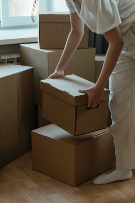 A person wearing a brown shirt is carefully unwrapping a large piece of white bubble wrap from a household item, likely during packing for a home relocation. The bubble wrap is partially unrolled and held with both hands, revealing the textured plastic surface designed for protecting furniture or appliances. The setting appears to be inside a well-lit room with wooden flooring, and visible background elements include a wooden bookshelf filled with books, a potted plant on a small stand, and a sheer white curtain allowing natural light to fill the space. This scene illustrates the careful packing process involved in home removals and furniture transport, highlighting the importance of secure wrapping during household moves coordinated by a professional service like Man With a Van Hillingdon, specializing in house removals and logistical moving solutions.
