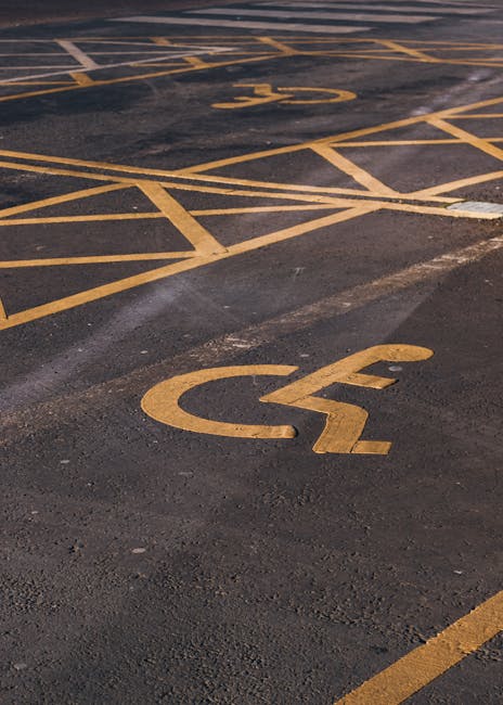 A section of an outdoor parking area showing a marked disabled parking space with yellow paint on dark asphalt. The space features a large wheelchair symbol painted on the ground, with additional yellow lines outlining the parking boundaries. The surface appears slightly worn, with some areas of faded paint and patches of dirt. In the background, parts of adjacent parking bays are visible, separated by yellow lines, and other marked parking spaces can be seen. The scene is well-lit, with natural light illuminating the markings clearly, providing a neutral, factual view suitable for accessibility purposes and relevant to parking arrangements in the context of house removals or moving services associated with Man With a Van Hillingdon.