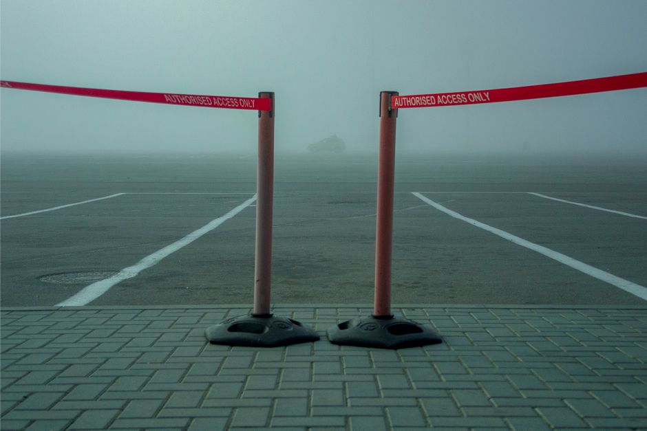 A foggy scene of a parking area with two temporary barriers made of pink poles and red tape marked 'Authorised Access Only' stretching between them, blocking the entrance to the empty parking spaces. The barriers are positioned on a paved area with interlocking bricks in the foreground, leading to the open asphalt surface with white line markings for parking bays. The thick fog limits visibility beyond the immediate area, with a faint outline of an object or distant structure barely visible in the background. This image relates to vehicle access control and parking management, which may be relevant for house removals or moving logistics in consultation with services such as Man With a Van Hillingdon, especially in the context of following local regulations regarding access during a home relocation or furniture transport.