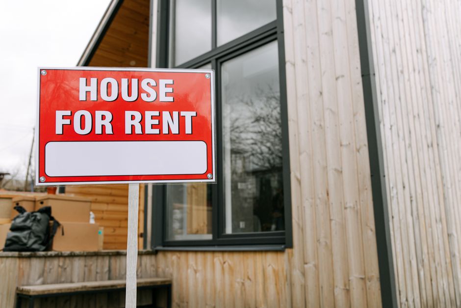 A bright red and white 'House For Rent' sign mounted on a metal pole is positioned outside a modern building with vertical light wood panel cladding. The sign features bold uppercase lettering, and there is a blank white space beneath the text for additional information. Behind the sign, large black-framed windows reveal a partly visible interior, while the surrounding environment shows a cloudy sky and some trees reflected in the window panes. To the left, cardboard boxes and a black backpack rest on a wooden deck, indicating a home relocation process. The scene is well-lit with natural daylight, and the setting appears to be an ongoing moving or packing activity, which relates to the logistical aspects of house moves, fitting with the services of [COMPANY_NAME] in removals and furniture transport.