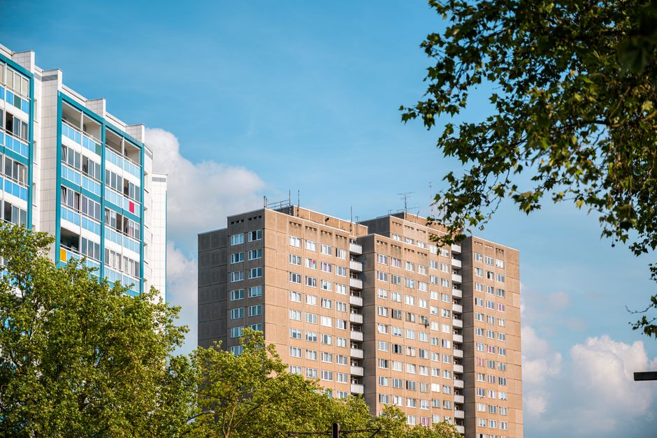 The image shows a high-rise residential building with a grey facade and multiple rows of windows, some of which have laundry or curtains visible. To the left, part of a modern blue and white apartment building with large glass balconies can be seen. In the foreground, lush green trees with dense foliage partially obscure the lower floors of the buildings, providing a natural contrast to the urban environment. The sky above is bright blue with some scattered white clouds, indicating a clear day. The scene captures a typical cityscape, which could be relevant to home relocation or moving services such as those offered by Man With a Van Hillingdon, especially when planning furniture transport or packing logistics for Hillingdon flats and complying with leasehold rules.