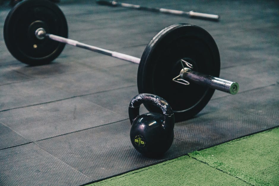 A black kettlebell and a barbell with black weight plates are placed on a rubber gym floor, with the kettlebell positioned in the foreground and the barbell extending into the background. The barbell, which appears to be in a loading process, has a metal bar with green end caps and is partially lifted or arranged for gym use. The gym floor consists of textured black mats, with a small section of green artificial turf near the bottom right corner. The scene is well-lit, emphasizing the equipment's metallic and rubber surfaces. This setup reflects typical home or commercial gym equipment, relevant to furniture transport and packing during moves, as handled by specialist removal services like Man With a Van Hillingdon, which manages safe relocation of various bulky items such as gym equipment.