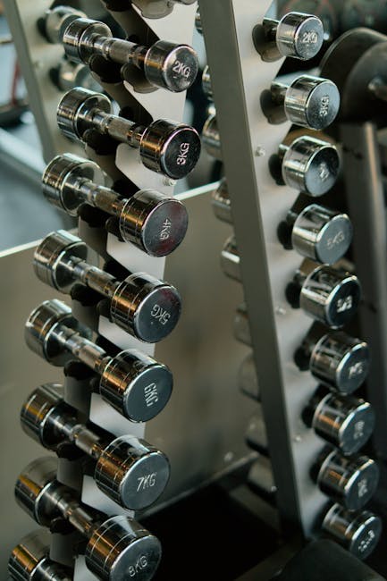 Close-up view of a set of black rubber-coated dumbbells arranged vertically on a metal rack inside a home gym or fitness space. The dumbbells are labeled with their weights in kilograms, ranging from 1kg to 7kg, with each weight clearly visible on the end caps. The rack is positioned near a doorway or opening, with some light illuminating the scene, highlighting the metallic reflections on the dumbbell handles. The background features a blurred indoor environment typical of a home workout area. This image reflects the preparation or packing of gym equipment that may be involved in a house removal or relocation service, illustrating the careful handling and organizational aspects of furniture and equipment transport facilitated by companies like Man With a Van Hillingdon.