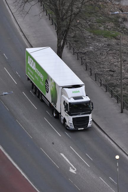 An aerial view of a white delivery truck with an ASDA Money advertising wrap on its trailer, parked on the side of a paved urban street with lane markings. The truck is positioned parallel to the sidewalk, which is lined with metal railings and a leafless tree. Adjacent to the pavement, there is a patch of bare ground with sparse vegetation. The truck appears to be used for home relocation or furniture transport as part of a moving service, possibly associated with house removals or packing and moving activities. The scene is captured during daylight with clear visibility, highlighting the vehicle’s size and positioning in the context of logistics for house removals, supported by the presence of loading equipment such as trolleys or straps if visible, though none are evident in this particular image.