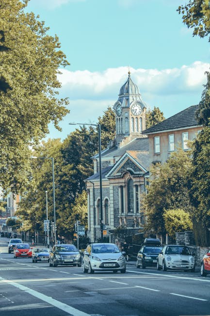 A street scene showing a busy parking area with multiple cars parked along the pavement, some vehicles queued in traffic on a clear day with blue sky and scattered clouds. To the right, there is a building partially obscured by leafy green trees, which extends towards a church with a prominent clock tower and a domed roof topped with a spire. The church features architectural details such as arched windows and decorative brickwork. Streetlights and traffic signals are visible along the road, indicating an urban environment typical of Hillingdon. The setting suggests an active residential or commercial neighbourhood, where home relocation or furniture transport services by Man With a Van Hillingdon might operate, with the scene capturing the typical transportation and loading environment during a city move or packing process.