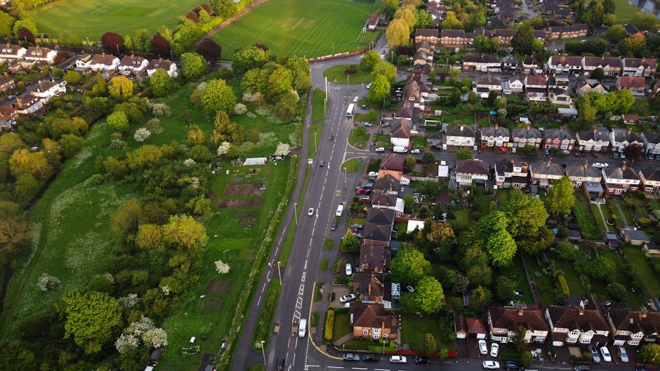 An aerial view of a residential neighbourhood illustrating a home relocation scene, with a narrow street lined with parked cars and houses on either side, surrounded by green trees and well-maintained gardens. In the foreground, the street is partially obstructed by a parked white van used for furniture transport, next to an opening in the pavement where a man with a van from Man With a Van Hillingdon is involved in a loading process, carrying cardboard boxes and wrapped furniture pieces toward the vehicle. The van is parked adjacent to a driveway, with some packing materials visible near the entrance of one of the homes, indicating ongoing packing and moving activities. The background features expansive green open spaces and more residential houses under a clear sky, emphasizing the suburban setting typical of the West Drayton to Yiewsley area, as part of a professional removals service supporting local house moves.