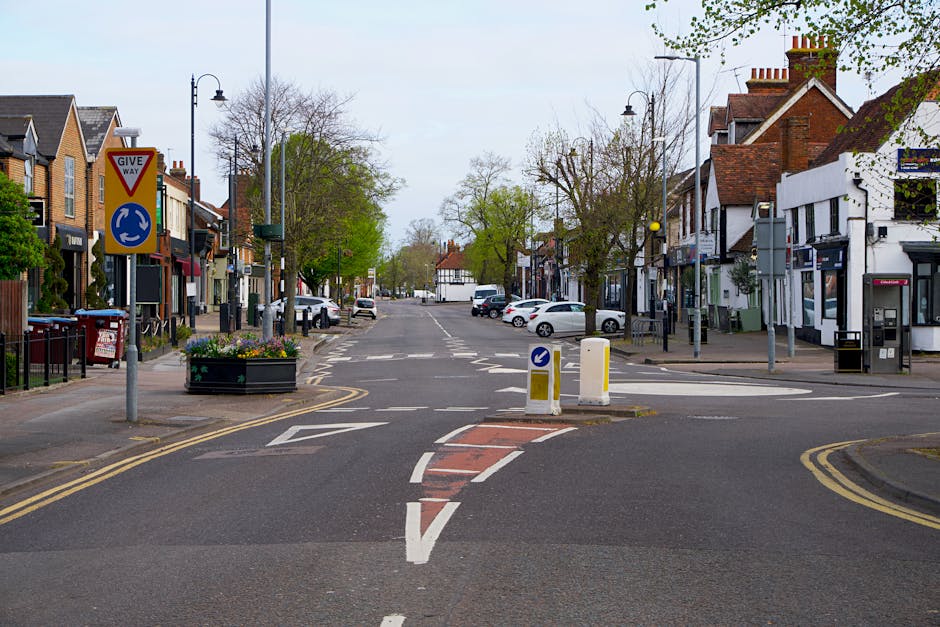 The image depicts a quiet residential street scene in West Drayton, with a clear view of the intersection leading into a town centre area. On the left side, there are several terraced houses and small shops with brick facades, some with decorative shop signage. Mature trees with green leaves line the pavement, and a large black planter filled with colorful flowers sits at the corner near a prominent give way sign and roundabout direction sign. The road is marked with white lines, including a temporary red and white paint section indicating ongoing or restricted vehicle movement, suitable for loading or home relocation purposes. Parked cars, including a white van, are visible along the street, with a few cars approaching or driving through the intersection. Tall streetlights and utility poles line the sidewalks, and the sky overhead appears overcast with diffuse lighting, typical of a calm day. This scene, associated with house removals and furniture transport, reflects typical urban logistics where vehicles such as [COMPANY_NAME]'s vans might load or unload furniture and boxes during a residential move, with clear traffic guidance ensuring safety and efficiency in the moving process.
