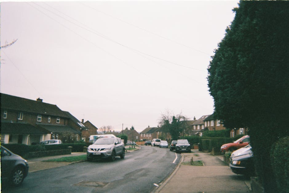 An aerial view of a residential neighbourhood illustrating a home relocation scene, with a narrow street lined with parked cars and houses on either side, surrounded by green trees and well-maintained gardens. In the foreground, the street is partially obstructed by a parked white van used for furniture transport, next to an opening in the pavement where a man with a van from Man With a Van Hillingdon is involved in a loading process, carrying cardboard boxes and wrapped furniture pieces toward the vehicle. The van is parked adjacent to a driveway, with some packing materials visible near the entrance of one of the homes, indicating ongoing packing and moving activities. The background features expansive green open spaces and more residential houses under a clear sky, emphasizing the suburban setting typical of the West Drayton to Yiewsley area, as part of a professional removals service supporting local house moves.
